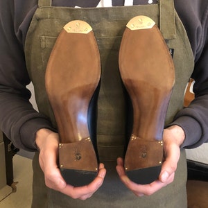 Puede incluir: Un zapatero sostiene dos zapatos de cuero negro, mostrando sus suelas. Las suelas son de color marr&oacute;n claro, con un tal&oacute;n y una puntera de color marr&oacute;n m&aacute;s oscuro. Cada tal&oacute;n tiene una peque&ntilde;a placa met&aacute;lica dorada. Los zapatos son nuevos y parecen hechos a mano.