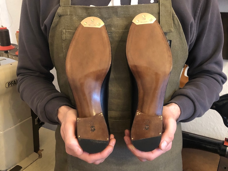 Puede incluir: Un zapatero sostiene dos zapatos de cuero negro, mostrando sus suelas. Las suelas son de color marr&oacute;n claro, con un tal&oacute;n y una puntera de color marr&oacute;n m&aacute;s oscuro. Cada tal&oacute;n tiene una peque&ntilde;a placa met&aacute;lica dorada. Los zapatos son nuevos y parecen hechos a mano.