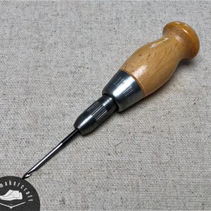 Puede incluir: Una lezna de zapatero con mango de madera clara y punta de metal plateado. La herramienta está diseñada para perforar cuero. Una etiqueta circular con texto y un gráfico de zapato es visible.
