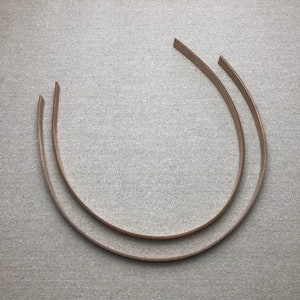 K&ouml;nnte beinhalten: Zwei hellbraune, gebogene Haarb&auml;nder liegen auf einer strukturierten, grauen Oberfl&auml;che. Die Haarb&auml;nder sind d&uuml;nn und scheinen aus Leder oder einem &auml;hnlichen Material zu bestehen. Die Enden der Haarb&auml;nder sind schr&auml;g geschnitten.