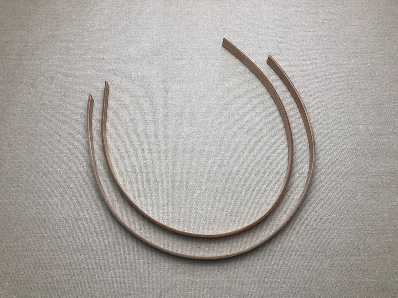 K&ouml;nnte beinhalten: Zwei hellbraune, gebogene Haarb&auml;nder liegen auf einer strukturierten, grauen Oberfl&auml;che. Die Haarb&auml;nder sind d&uuml;nn und scheinen aus Leder oder einem &auml;hnlichen Material zu bestehen. Die Enden der Haarb&auml;nder sind schr&auml;g geschnitten.