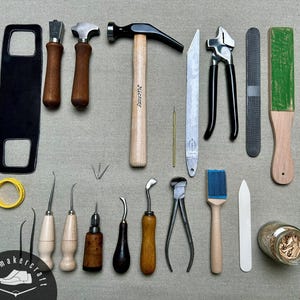 Può includere: Una collezione di attrezzi da calzolaio che include un martello con manico in legno, vari punteruoli con manici in legno, pinze in metallo, una raspa, un coramella verde, un rotolo di nastro giallo, una spazzola, una stecca d'osso e un barattolo di vetro.