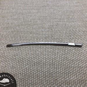 May include: A curved, silver-colored shoemaking tool, possibly a needle or awl, lies on a textured gray surface. The tool has a slightly curved shape with a blunt end and a small, rectangular white piece near the other end.