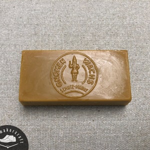 Puede incluir: Una barra rectangular de cera para zapatos de color marrón claro. La parte superior presenta las palabras "GNOMEN WACHS" y un logotipo de gnomo. La cera está sobre una superficie texturizada de color gris claro.