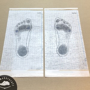 May include: Two foot impression tracings on graph paper. Each tracing shows the outline of a foot with a dark gray ink fill. The paper has a grid pattern and is labeled with handwritten numbers. The shoemaker logo is in the bottom left corner.