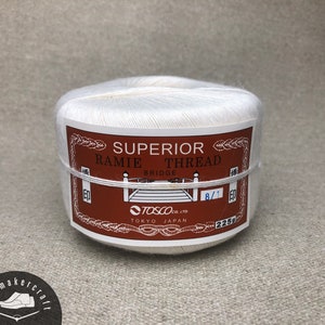 Puede incluir: Un carrete de hilo de ramio blanco superior, etiquetado con texto marrón y blanco. La etiqueta incluye el texto "BRIDGE" y "TOSCO CO. LTD TOKYO JAPAN". El carrete está envuelto con una banda fina y transparente.