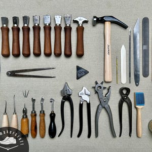 Könnte beinhalten: Eine Sammlung von Lederwerkzeugen, darunter ein Hammer mit Hickory-Stiel, verschiedene Metallahlen mit Holzgriffen, Zangen, eine Revolverlochzange, ein Abziehriemen mit grüner Polierpaste, ein Metallzirkel und ein kleines Glas mit Spänen.