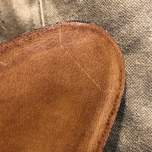 Puede incluir: Primer plano de una secci&oacute;n de cuero marr&oacute;n, posiblemente de un zapato o bolso. El cuero tiene una superficie texturizada con grano visible y ligeras imperfecciones. El borde est&aacute; cosido con hilo oscuro, y el fondo es una tela texturizada.