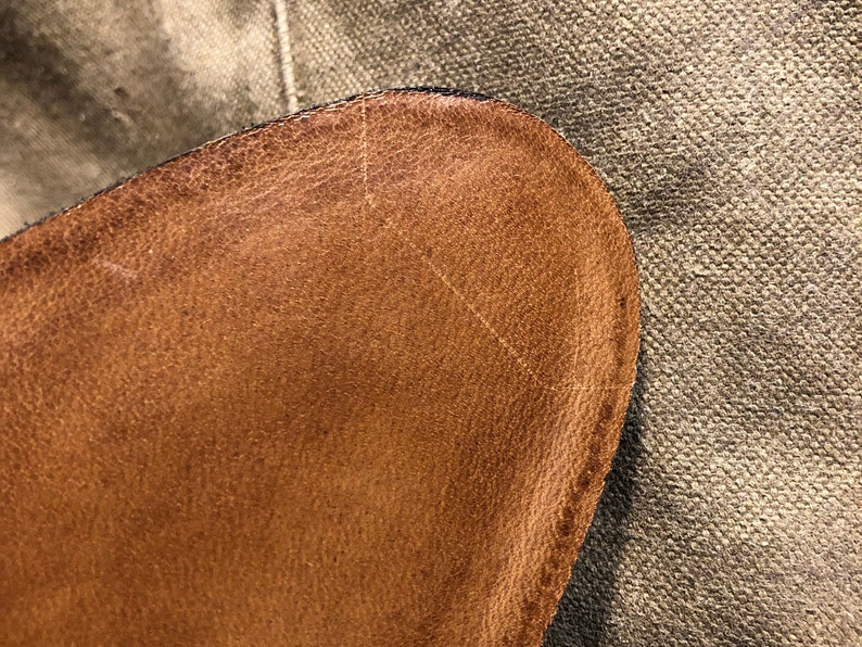 Puede incluir: Primer plano de una secci&oacute;n de cuero marr&oacute;n, posiblemente de un zapato o bolso. El cuero tiene una superficie texturizada con grano visible y ligeras imperfecciones. El borde est&aacute; cosido con hilo oscuro, y el fondo es una tela texturizada.