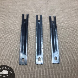 Pode incluir: Três suportes de metal pretos com base retangular e topo em forma de U. Cada suporte tem um furo perto da base e outro perto do topo. Os suportes estão dispostos verticalmente sobre uma superfície bege texturizada.
