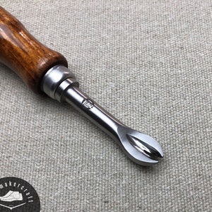 Könnte beinhalten: Ein Schuhmacherwerkzeug mit Holzgriff und Metallkopf. Der Griff ist warmbraun, und der Metallkopf hat ein gebogenes, spitzes Ende. Das Werkzeug ist für die Lederverarbeitung bestimmt und trägt den Text "7E" auf dem Metall.