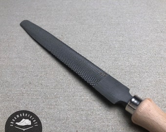 Könnte beinhalten: Eine Raspel mit einer strukturierten, dunkelgrauen Oberfläche und einem Holzgriff. Die Raspel dient zum Formen und Glätten von Materialien. Der Griff ist helles Holz mit einer Metallzwinge. Das Werkzeug wird wahrscheinlich in der Holzbearbeitung oder Schuhmacherei verwendet.