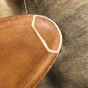 Puede incluir: Primer plano de un objeto de cuero marr&oacute;n con un dise&ntilde;o geom&eacute;trico. El objeto tiene un acento de color beige claro y un borde oscuro. El cuero parece texturizado y desgastado, lo que sugiere un art&iacute;culo vintage o artesanal.