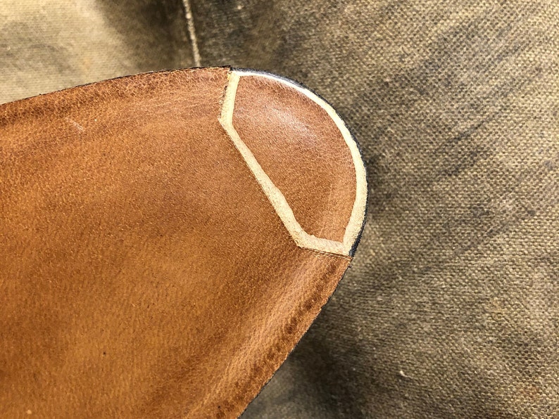 Puede incluir: Primer plano de un objeto de cuero marr&oacute;n con un dise&ntilde;o geom&eacute;trico. El objeto tiene un acento de color beige claro y un borde oscuro. El cuero parece texturizado y desgastado, lo que sugiere un art&iacute;culo vintage o artesanal.