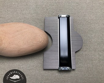 Könnte beinhalten: Ein Holzschuhleisten neben einer Metallkonturenlehre. Die Lehre hat eine schwarze Mitte und ist mit silbernen Nieten befestigt. Das Logo des Schuhmachers befindet sich in der unteren linken Ecke.
