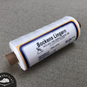 Puede incluir: Una bobina blanca de hilo de lino Bockens Lingarn, con una etiqueta con texto y un logotipo. La etiqueta tiene acentos azules y amarillos. La bobina está sobre una superficie de color neutro. El texto de la etiqueta incluye "HOLMA-HELSINGLANDS AB" y "100% Lin/Flax".