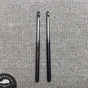 Puede incluir: Dos agujas de ganchillo plateadas con puntas oscuras se muestran sobre un fondo de tela gris texturizada. Las agujas están colocadas verticalmente, una al lado de la otra. La imagen incluye un logotipo circular en la esquina inferior izquierda. Las agujas son nuevas.