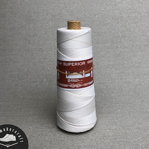 Puede incluir: Una bobina de hilo blanco con la etiqueta "SUPERIOR RAMIE THREAD BRIDGE" con una etiqueta marrón y una tapa de corcho. La etiqueta también incluye el texto "TOSCO CO., LTD. TOKYO JAPAN". El hilo está bien enrollado.