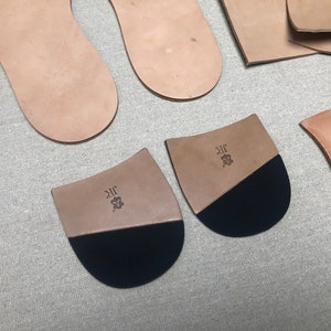 K&ouml;nnte beinhalten: Verschiedene Lederschuhkomponenten, darunter Innensohlen und Fersenteile. Die Fersenteile sind zweifarbig, mit einem Oberteil aus Naturleder und einem unteren Teil aus schwarzem Gummi. Die Initialen "JR" und ein kleines Emblem sind auf das Leder gestempelt.