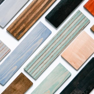 Op de afbeelding: Een assortiment rechthoekige houten planken in diverse kleuren, waaronder bruin, zwart, blauw en wit. Sommige planken hebben een gestreept patroon, terwijl andere effen zijn. De planken zijn diagonaal gerangschikt tegen een witte achtergrond, waarbij de verschillende houtnerven en afwerkingen worden getoond.