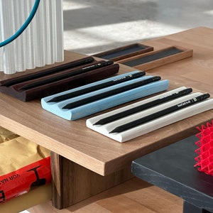 May include: Three pencil holders made of ceramic in brown, light blue, and white, each holding two black pencils. The holders are on a wooden surface.