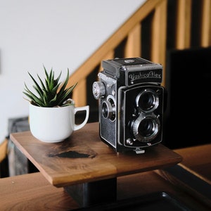 Peut inclure: Un appareil photo vintage Yashica Flex noir et argenté posé sur une étagère en bois avec une petite plante succulente en pot. L'appareil photo a deux objectifs et un bouton argenté sur le côté.
