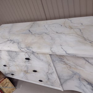 May include: A white dresser with a marble-like finish. The dresser has three drawers with black handles. The top of the dresser is a flat surface with a marble-like finish.