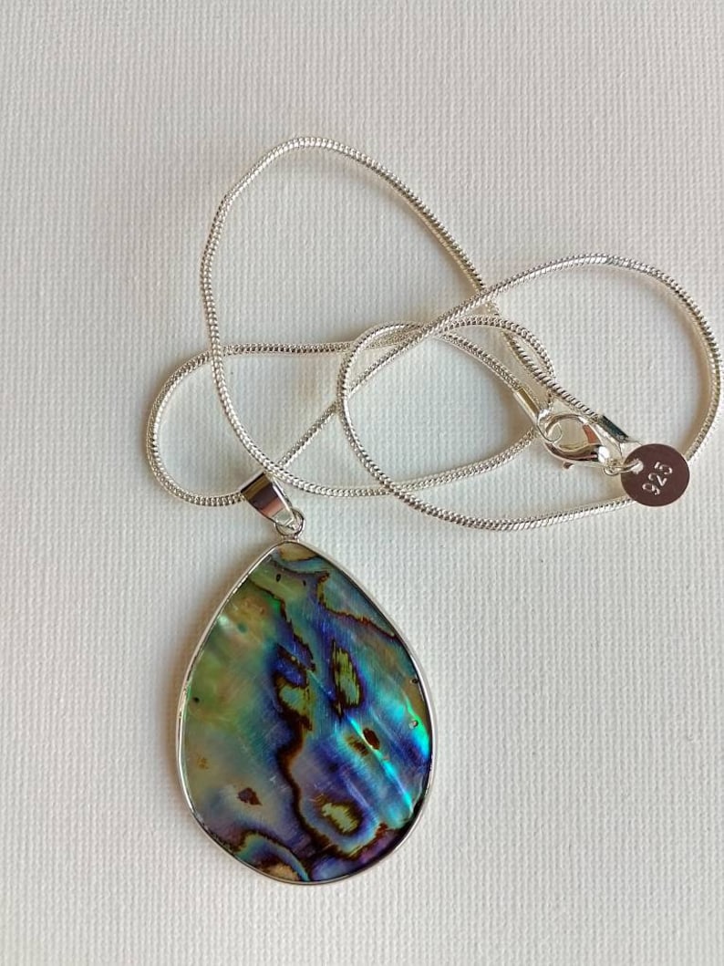 Puede incluir: Un collar de cadena de plata con un colgante de concha de abul&oacute;n en forma de l&aacute;grima. La concha tiene colores iridiscentes verdes, azules y morados.