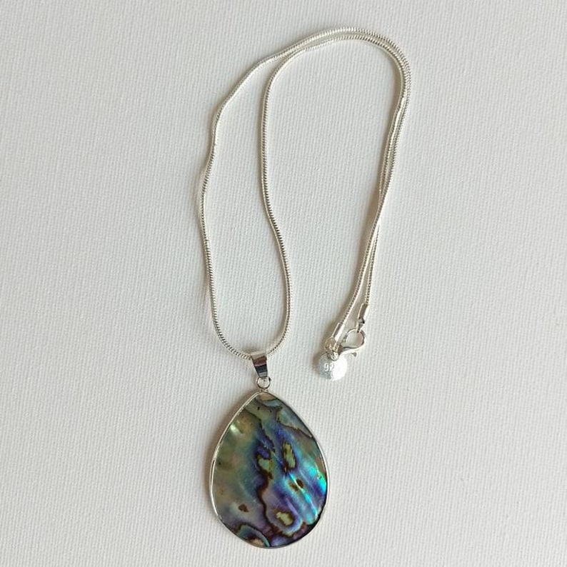Puede incluir: Un collar de cadena de plata con un colgante en forma de l&aacute;grima hecho de concha de abul&oacute;n iridiscente. El colgante tiene tonos de verde, azul y morado.
