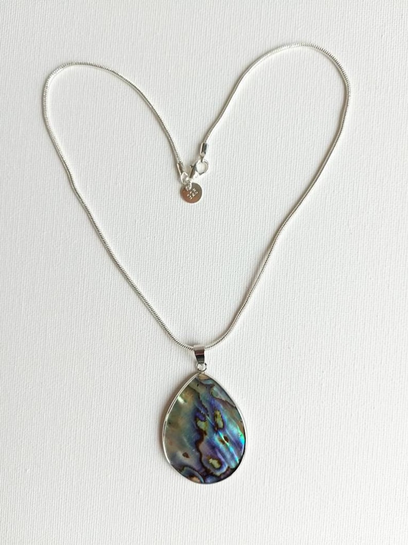 Puede incluir: Un collar de cadena de plata con un colgante de concha de abul&oacute;n en forma de l&aacute;grima. La concha tiene colores iridiscentes azules, verdes y morados.
