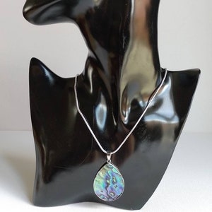 Puede incluir: Un collar de cadena de plata con un colgante en forma de l&aacute;grima hecho de concha de abul&oacute;n iridiscente. El colgante tiene tonos de verde, azul y morado.