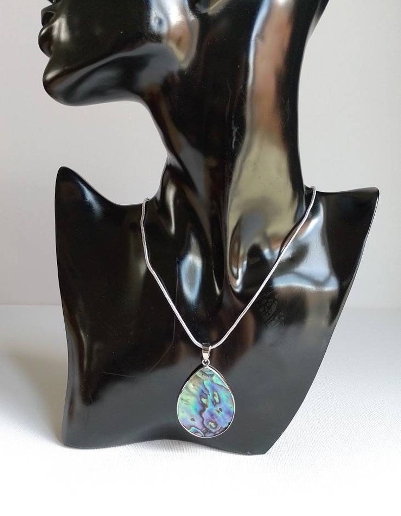 Puede incluir: Un collar de cadena de plata con un colgante en forma de l&aacute;grima hecho de concha de abul&oacute;n iridiscente. El colgante tiene tonos de verde, azul y morado.