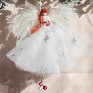 May include: A hanging angel ornament featuring a figure with long orange hair, white feathered wings, and a white tulle dress adorned with silver glitter stars. The figure wears a small pink rose at the waist and has pink rose accents on the feet.