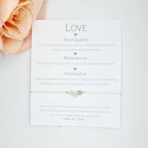 May include: A white card with the word "LOVE" in large, grey letters. The card lists the properties of three gemstones: Rose Quartz, Moonstone, and Aventurine. A bracelet with three beads, pink, green, and white, is tied to the card.
