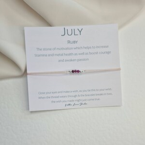 May include: A pink string bracelet with a small card attached. The card says "JULY RUBY" and has text about the stone of motivation. The bracelet has small silver beads and four small ruby-coloured beads.