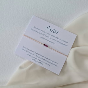 May include: A dainty bracelet with small, bright red ruby beads and silver details on a beige cord. The bracelet is presented on a card with the word "RUBY" and text about its properties.