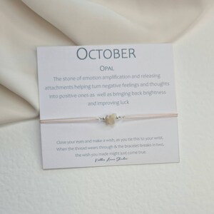 May include: A pink string bracelet with a small opal stone charm. The card attached to the bracelet reads "OCTOBER OPAL The stone of emotion amplification and releasing attachments helping turn negative feelings and thoughts into positive ones as well as bringing back brightness and improving luck Close your eyes and make a wish, as you tie this to your wrist. When the thread wears through & the bracelet breaks in two, the wish you made might just come true. Nellier Anne Studio"