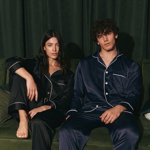 K&ouml;nnte beinhalten: Ein Paar tr&auml;gt passende schwarze und marineblaue Satin-Pyjamas und sitzt auf einem gr&uuml;nen Samtsofa. Die Frau tr&auml;gt ein schwarzes Pyjama-Oberteil mit wei&szlig;em Besatz und der Mann tr&auml;gt ein marineblaues Pyjama-Oberteil mit wei&szlig;em Besatz.