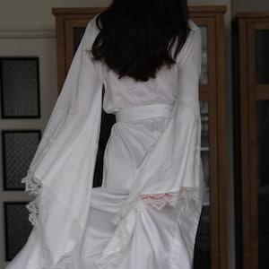 May include: A white satin robe with lace trim and long sleeves. The robe is being worn by a person standing on a white bed.
