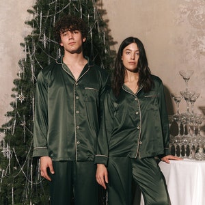 May include: Two people in matching dark green satin pyjamas with light pink trim, posing in front of a Christmas tree. The pyjamas consist of a button-down shirt and trousers. A table with champagne glasses and a bottle of champagne is in the background.