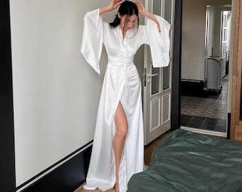 Può includere: Una vestaglia bianca in raso con maniche lunghe e chiusura avvolgente. La vestaglia ha uno spacco sul davanti e una cintura in vita. La vestaglia è indossata da una persona in una camera da letto.