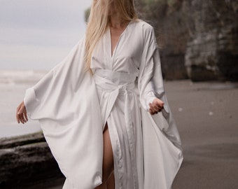 Può includere: Una veste bianca fluida in stile kimono con maniche lunghe e una cintura annodata in vita. La veste è indossata da una persona in piedi su una spiaggia.