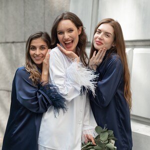 May include: Three women wearing matching pyjamas. The woman in the centre is wearing a white pyjama top with white feather trim and white trousers. The women on either side are wearing navy blue pyjama tops and trousers with feather trim. The woman in the centre is holding a bouquet of greenery.