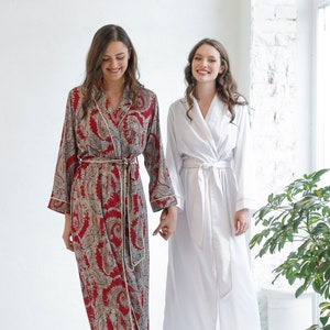 May include: Two women wearing long robes, one in a white robe and the other in a red and blue paisley robe. Both women are wearing pink slippers.
