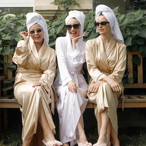 May include: Three women wearing white and gold bathrobes and sunglasses are sitting on a wooden bench. They are all wearing pink fluffy slippers with bows.