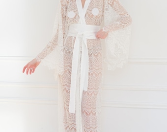May include: An ivory white lace robe featuring long, wide sleeves, a smooth satin-like collar, and a matching wide fabric tie belt at the waist. The garment is crafted from intricate floral lace with scalloped edges and includes the text Ivory White.