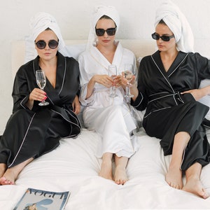 May include: Three women wearing white towels on their heads and black satin robes with white piping. They are sitting on a white bed with glasses of champagne in their hands. The text "Classy Black" is at the bottom of the image.