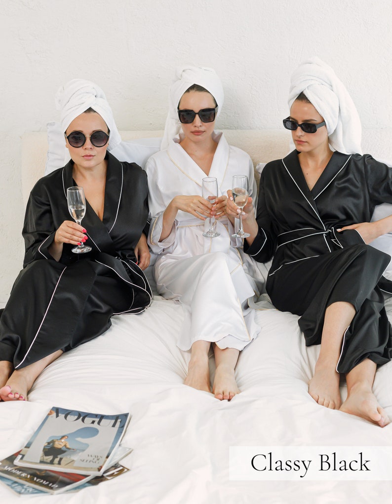 May include: Three women wearing white towels on their heads and black satin robes with white piping. They are sitting on a white bed with glasses of champagne in their hands. The text "Classy Black" is at the bottom of the image.
