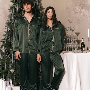 K&ouml;nnte beinhalten: Ein Paar tr&auml;gt passende dunkelgr&uuml;ne Satin-Pyjamas und steht vor einem Weihnachtsbaum. Der Mann steht links und die Frau rechts. Beide sind barfu&szlig;.
