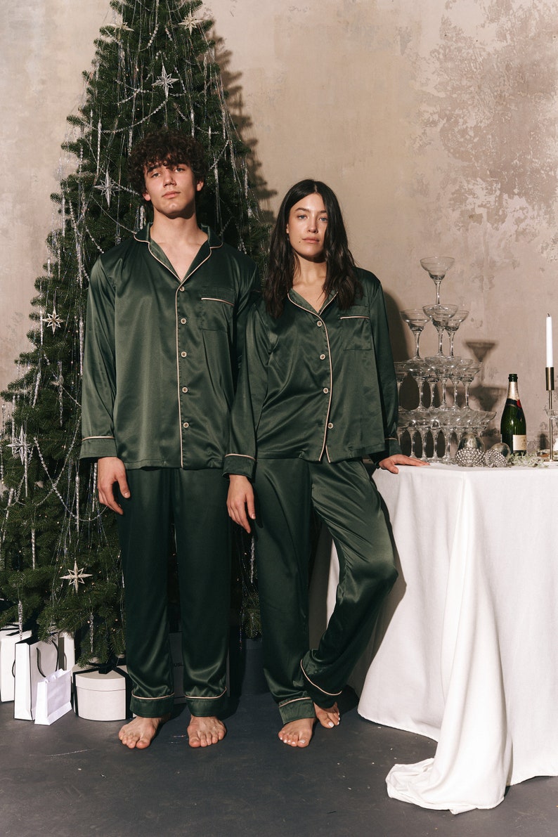K&ouml;nnte beinhalten: Ein Paar tr&auml;gt passende dunkelgr&uuml;ne Satin-Pyjamas und steht vor einem Weihnachtsbaum. Der Mann steht links und die Frau rechts. Beide sind barfu&szlig;.